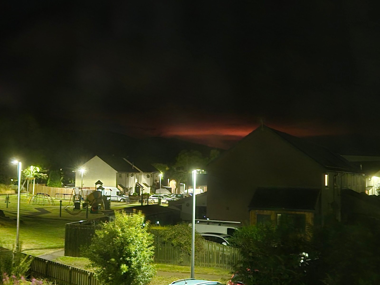Night-time view from Aviemore during the 2026 Cairngorm wildfire, with houses in the foreground and an orange glow from the fire visible beyond the dark hillside.