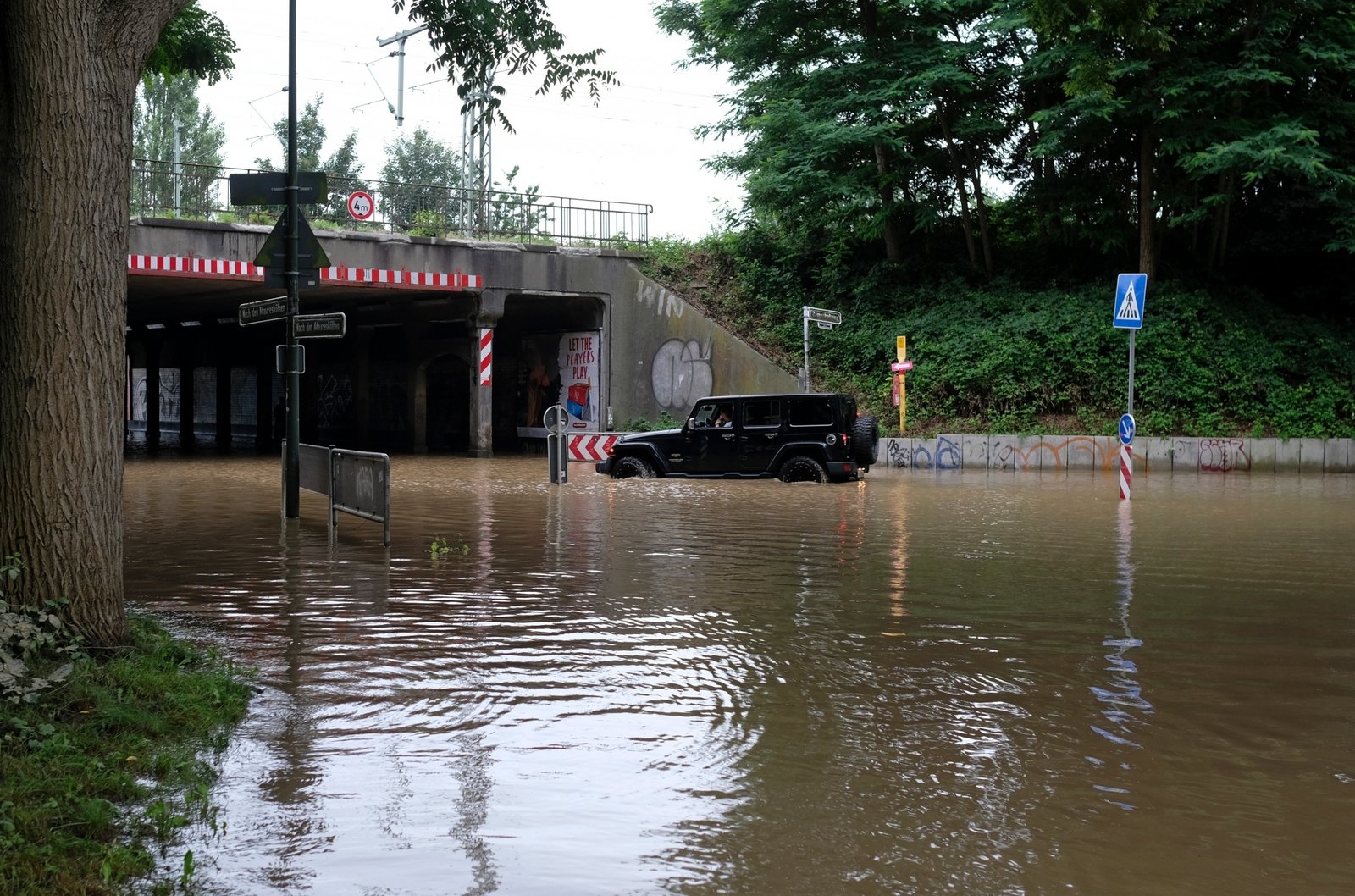 Urban street flooded with deep water around a road underpass, with a vehicle attempting to pass through.